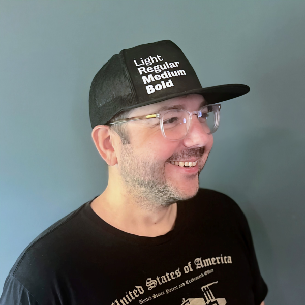 Portrait of Phil Carey-Bergren shot at an angle. Wearing a black and white hat with text, "Light, Regular, Medium, Bold" and a t-shirt for the US Patent and Trademark Office.